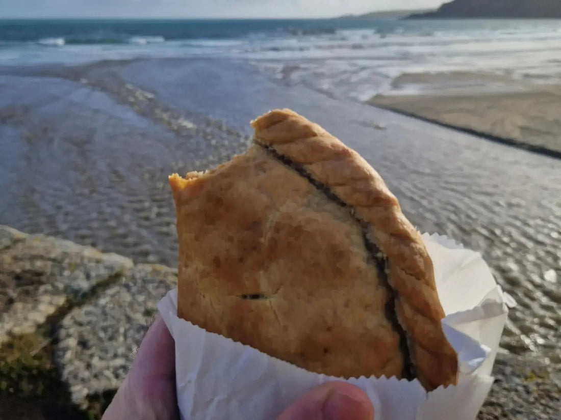 Vegan pasty in a white greaseproof bag held up against a beachy coastal backdrop